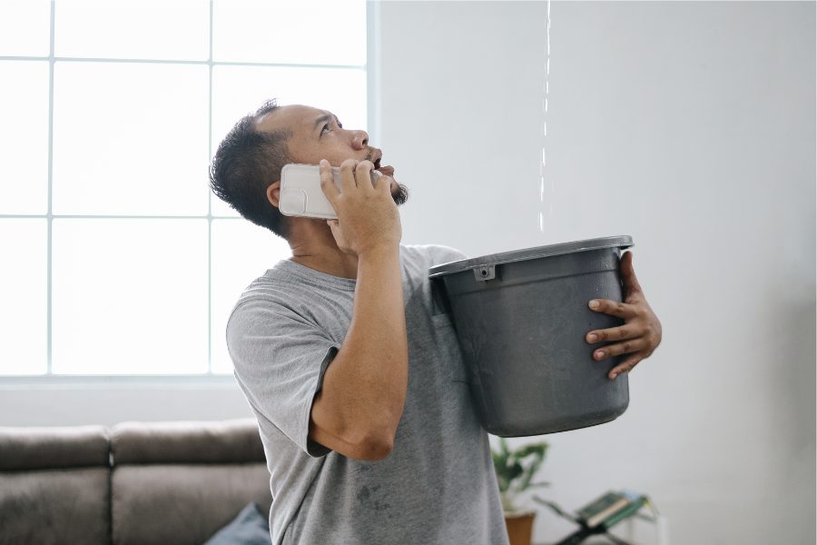A man holds a bucket under a leak in his ceiling while on the phone calling for help.