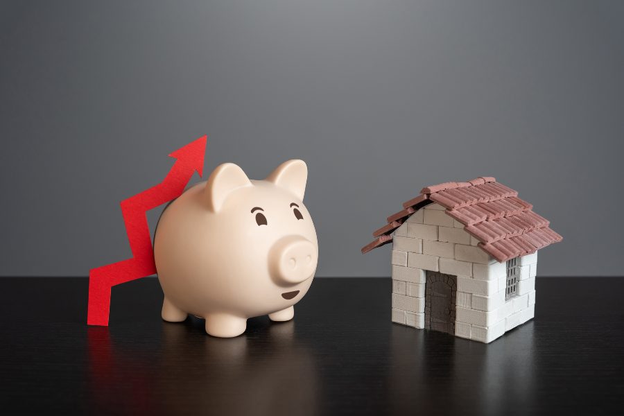 A piggy bank with a red up arrow leaning against it beside a small model home on a black table.