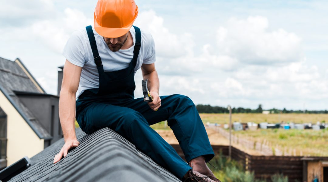 Roof inspection checklist B5 hero A roofer in blue coveralls, a white t-shirt, and orange hard hat sits in the peak of an asphalt shingle roof, inspecting the shingles.