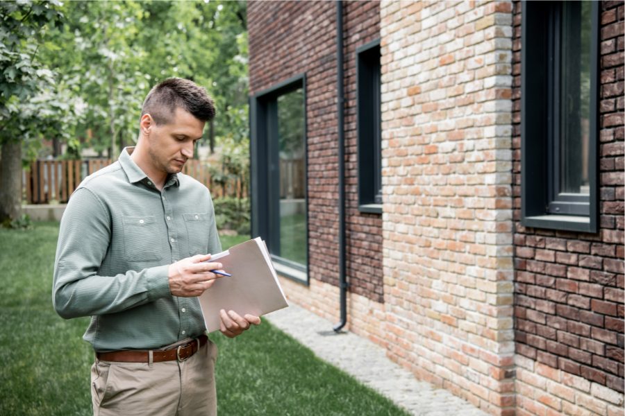 A homeowner wearing beige pants and a green button-up checks the exterior of his home, carrying a notepad and pen