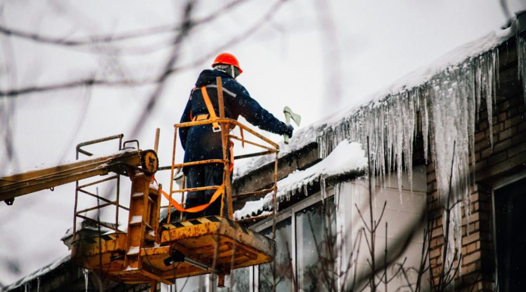 A roofer on a yellow mechanical lift removes thick sheets of ice from a roof with a small axe.